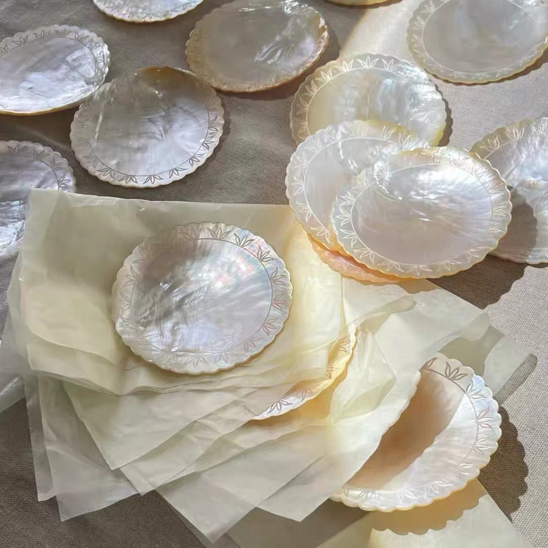 Collection of iridescent mother-of-pearl scallop shell dishes with engraved leaf border detail, individually wrapped in tissue paper and displayed on linen fabric in warm sunlight