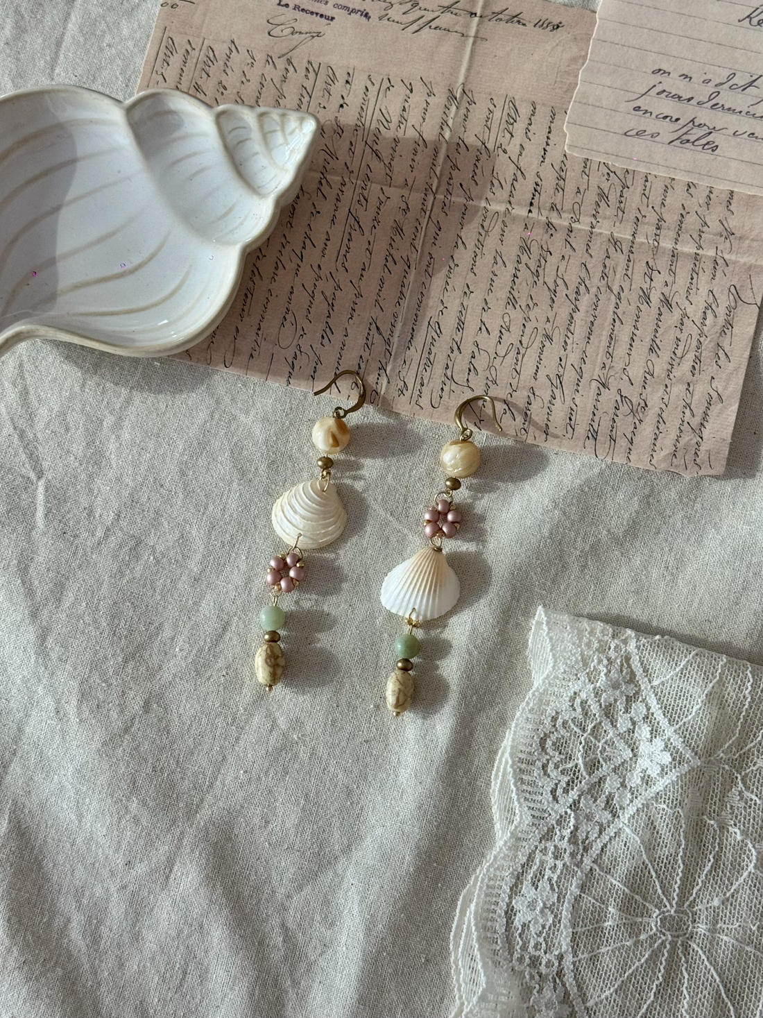 Handmade shell and bead drop earrings with pink flower accents and gold hardware, styled on linen fabric with vintage script letters and white lace