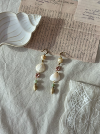 Handmade shell and bead drop earrings with pink flower accents and gold hardware, styled on linen fabric with vintage script letters and white lace
