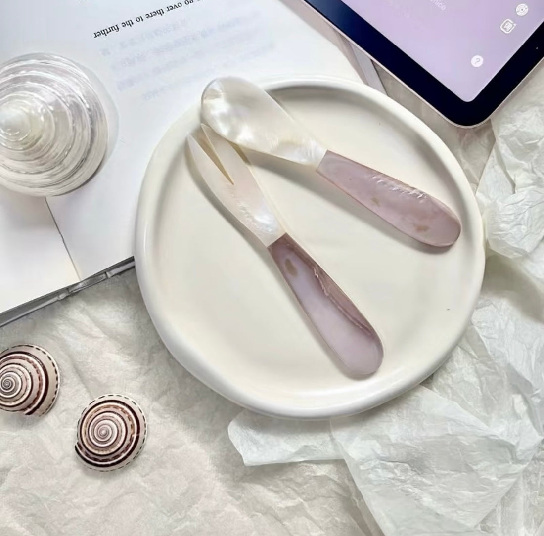 Pearl shell spoon and pink crystal spatula on a white ceramic plate, styled with spiral shells and a glass on white tissue paper