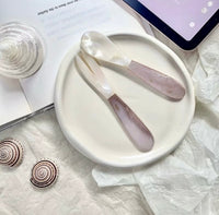 Pearl shell spoon and pink crystal spatula on a white ceramic plate, styled with spiral shells and a glass on white tissue paper