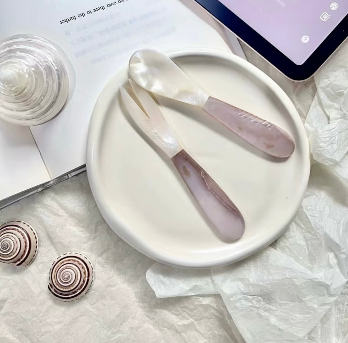 Pearl shell spoon and pink crystal spatula on a white ceramic plate, styled with spiral shells and a glass on white tissue paper