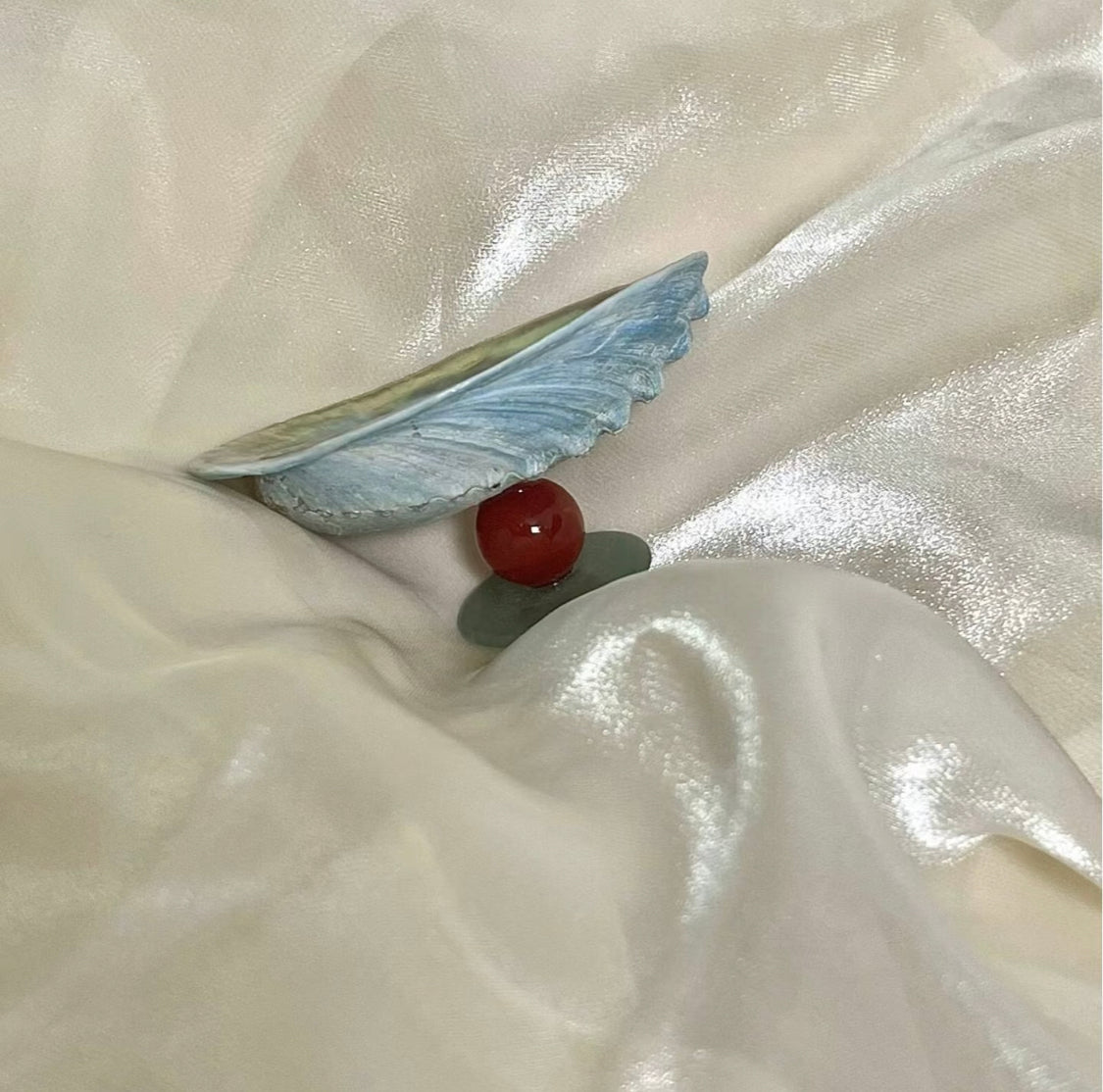 Blue iridescent mussel shell resting on shimmery white satin fabric alongside a deep red carnelian bead and a small green jade stone, styled as a natural wellness flat lay