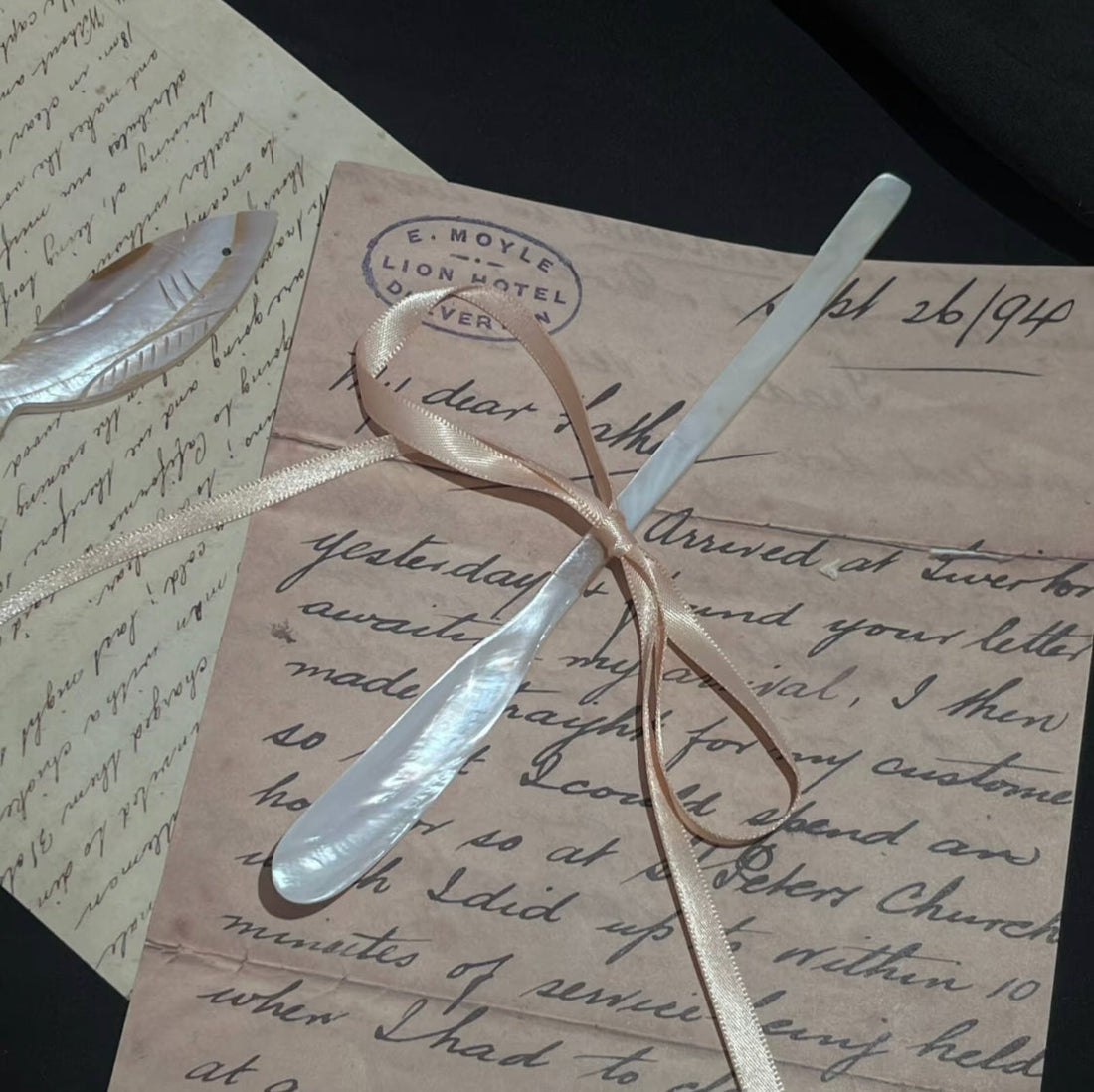 Mother-of-pearl butter spreader and shell fish brooch resting on vintage handwritten letters tied with a satin ribbon, stamped with E. Moyle Lion Hotel postmark on black background