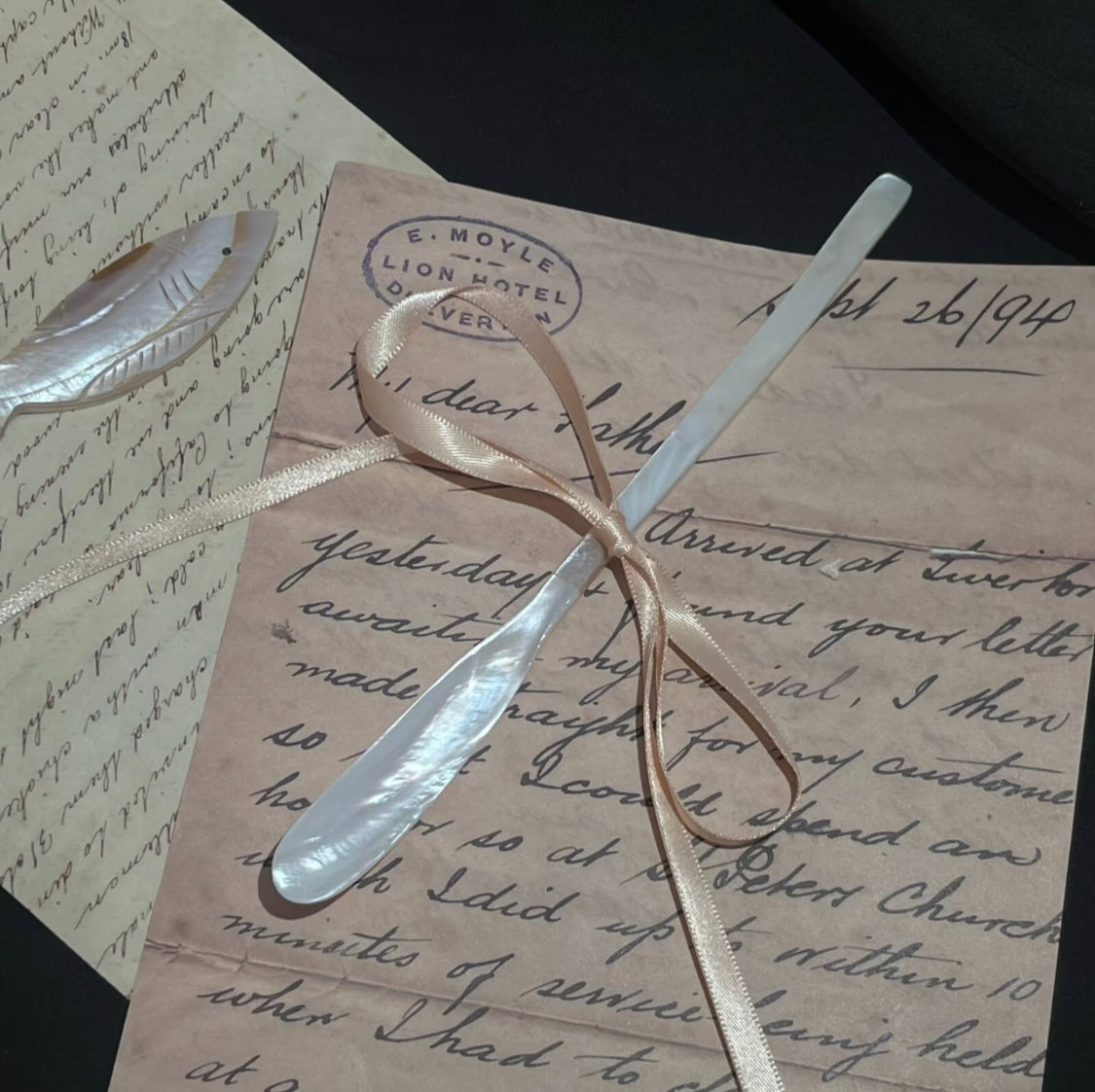 Mother-of-pearl butter spreader and shell fish brooch resting on vintage handwritten letters tied with a satin ribbon, stamped with E. Moyle Lion Hotel postmark on black background