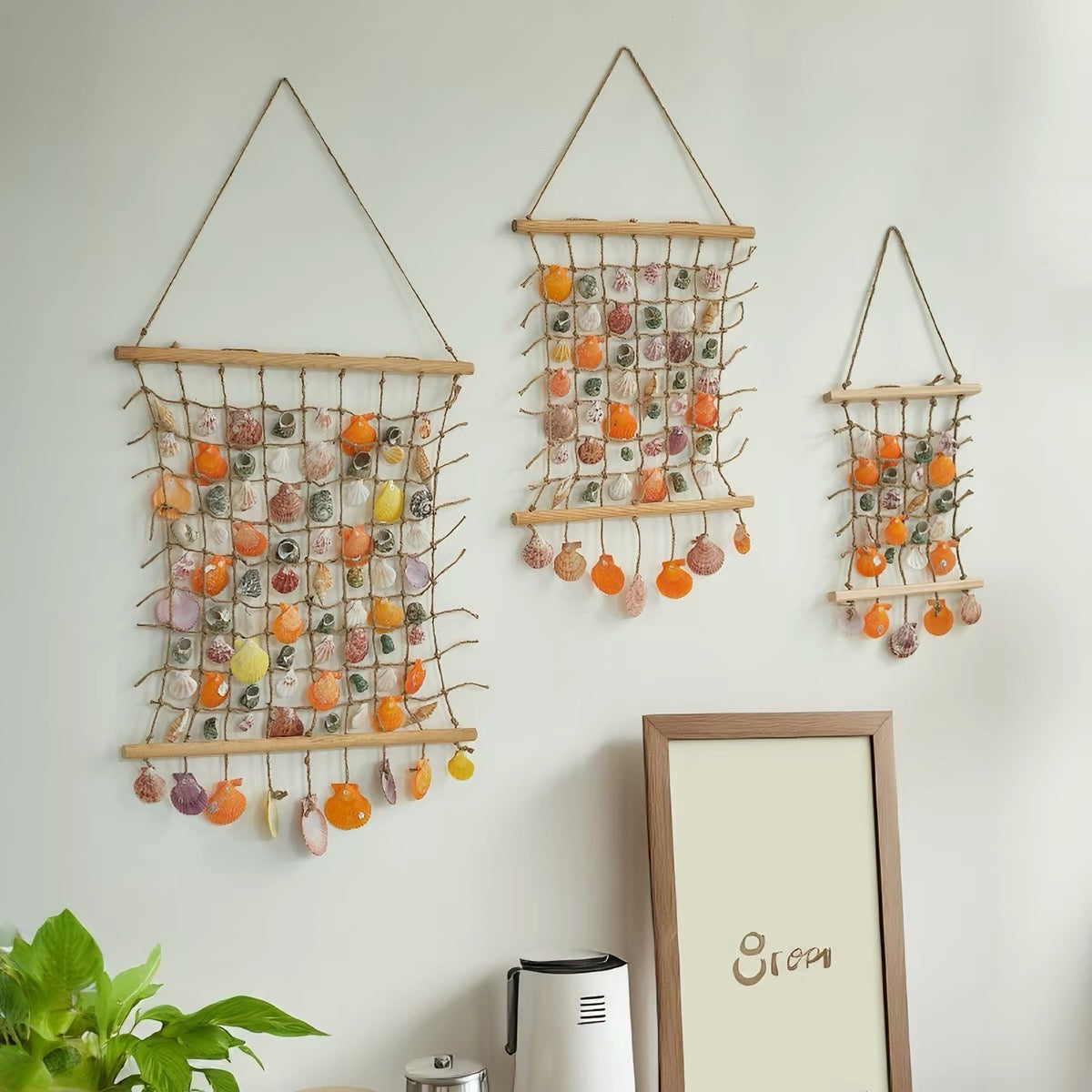 Three handmade shell wall hangings in different sizes made of jute rope net and wooden dowels, decorated with colorful orange, pink and green shells, displayed on a white wall