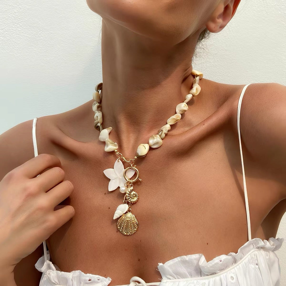 Woman wearing a natural cowrie shell necklace with white flower, gold scallop shell pendant and pearl charms on a gold toggle clasp, paired with a white ruffle top