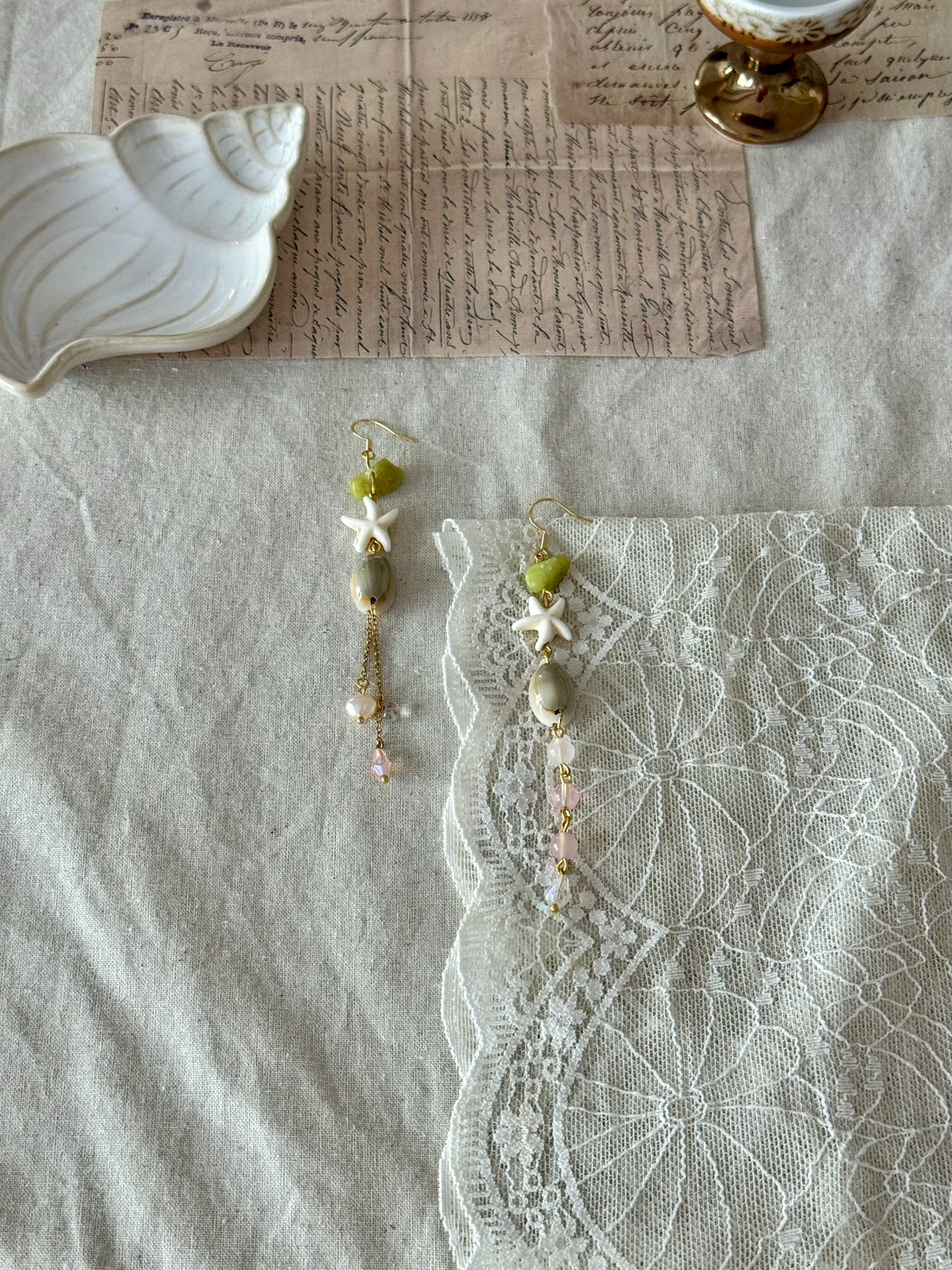 Handmade starfish and jade bead drop earrings with pink crystal and gold chain accents, styled on linen fabric with white lace, vintage script paper and a ceramic shell dish