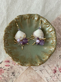 Handmade shell earrings with amethyst purple beads, crystal accents and gold spike details displayed on a green ceramic dish with floral paper background