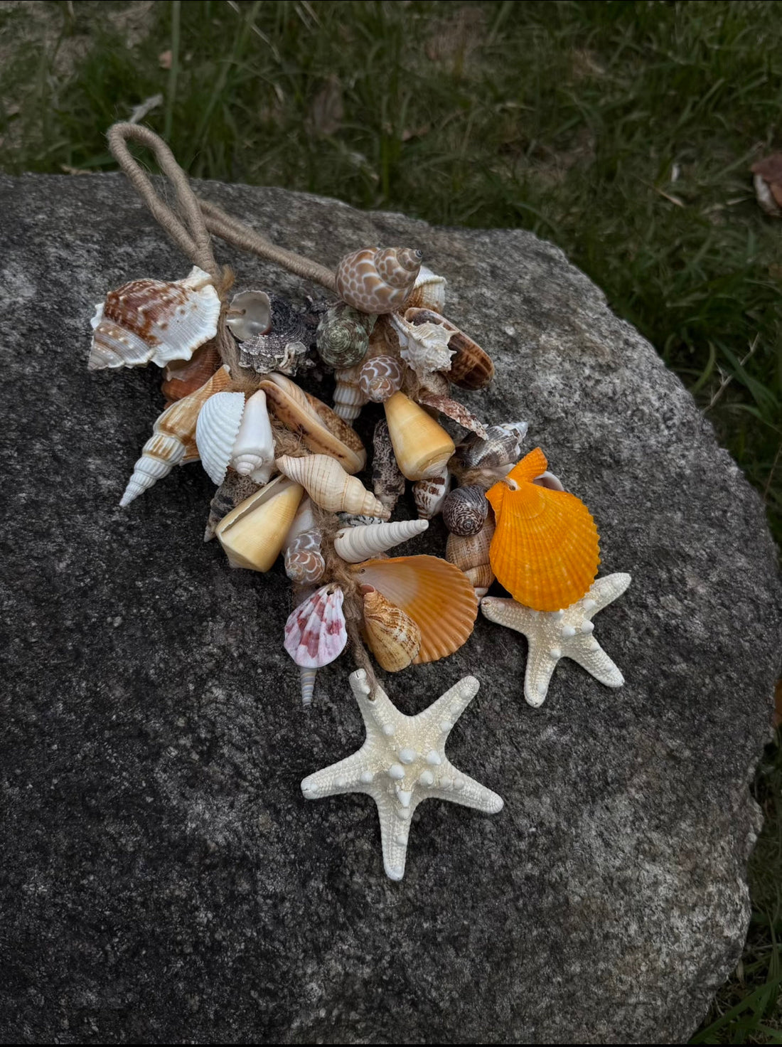 Handmade coastal shell cluster ornament on a granite rock featuring orange scallop shells, spiral cone shells, white starfish, and a silver turtle charm tied with jute twine