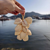 Hand holding a bunch of cream-colored spotted clam shells bundled together with jute twine against a scenic harbor and mountain backdrop in golden sunlight